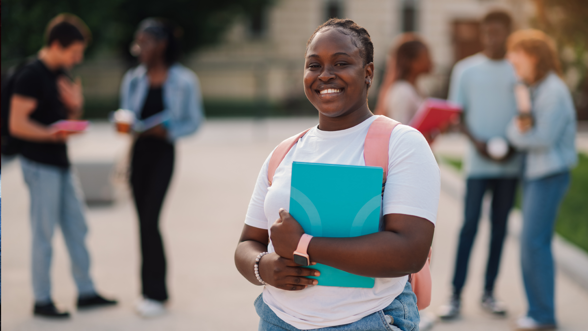 Smiling university student carrying a backpack and holding a notebook while standing on campus, with other students socialising and studying in the background.