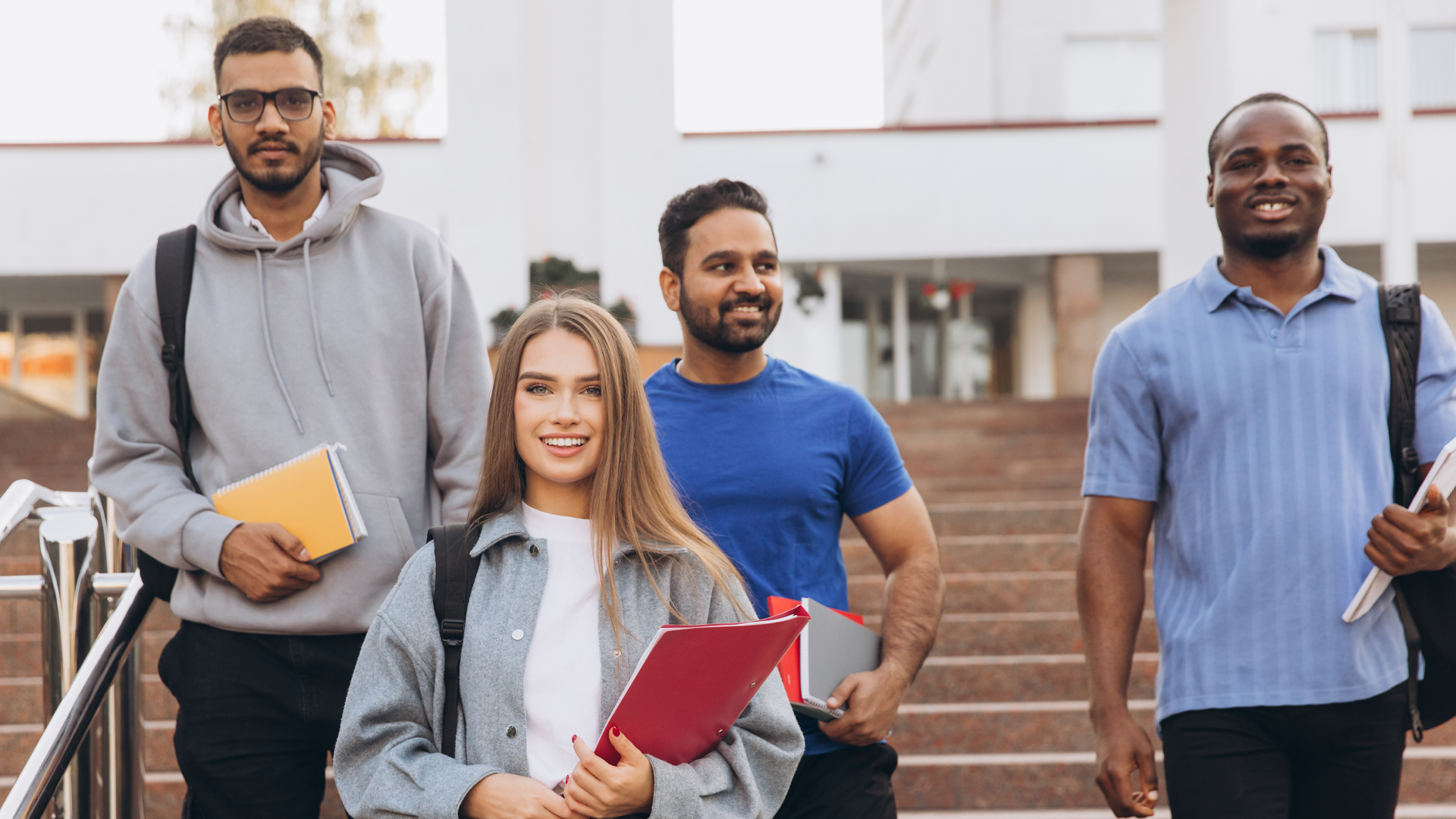 Four postsecondary students of diverse backgrounds walk together down campus steps carrying notebooks and backpacks.