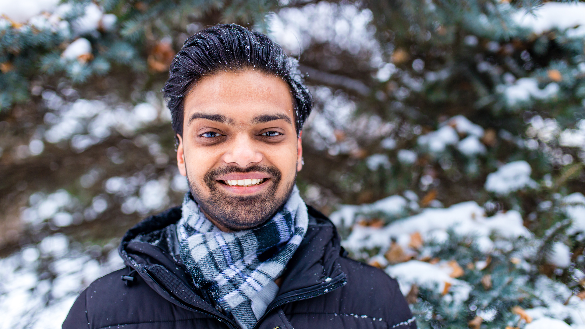 A smiling international student dressed for winter stands outdoors in front of snow-covered evergreen trees, wearing a black winter jacket and plaid scarf with snowflakes resting on their hair and shoulders.