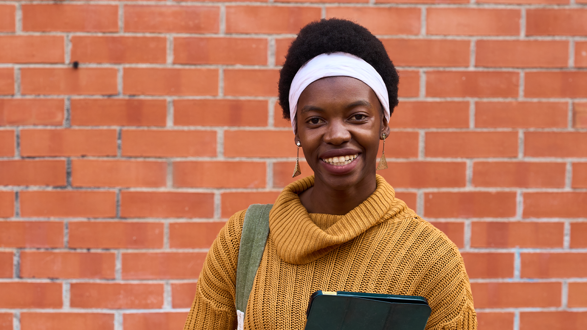 A smiling international student wearing a mustard knit sweater and white headband stands in front of a red brick wall, carrying a backpack and holding a tablet while looking directly at the camera.