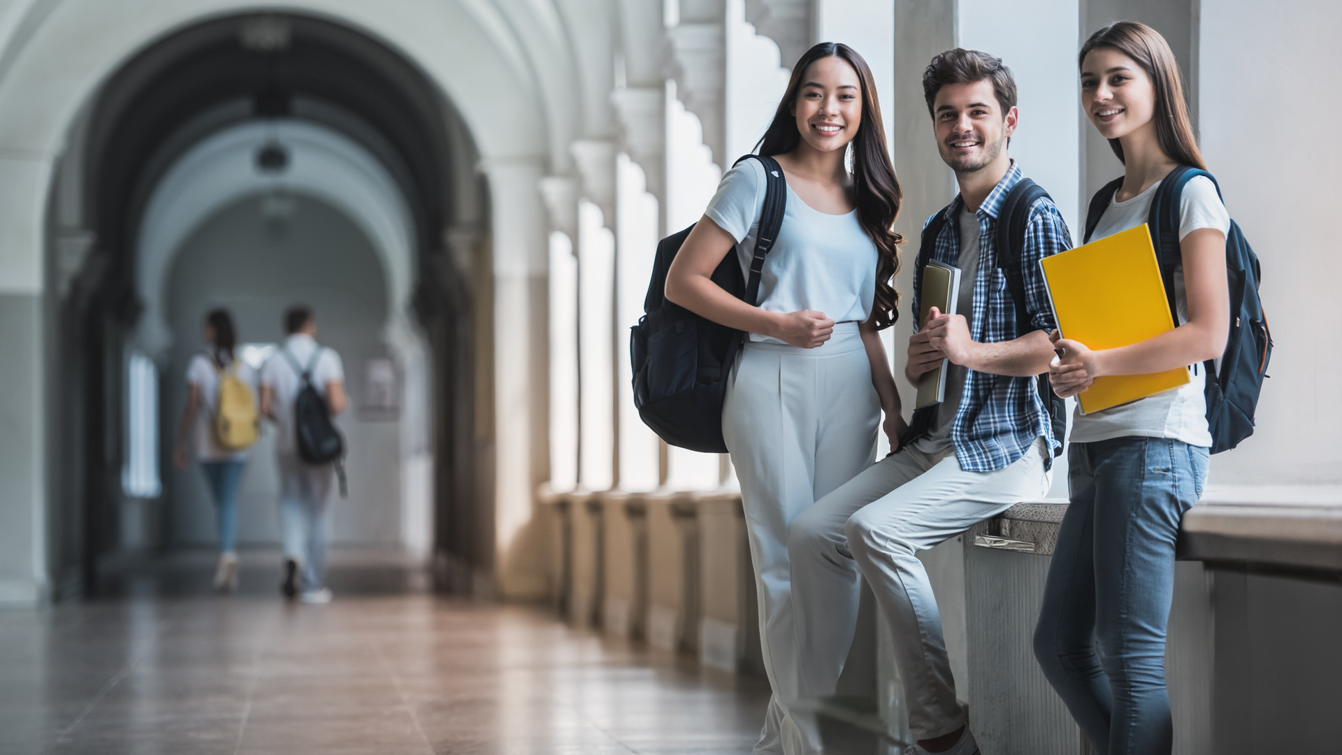 Three university students with backpacks smiling in a bright campus corridor, representing student success, engagement, and retention in higher education.